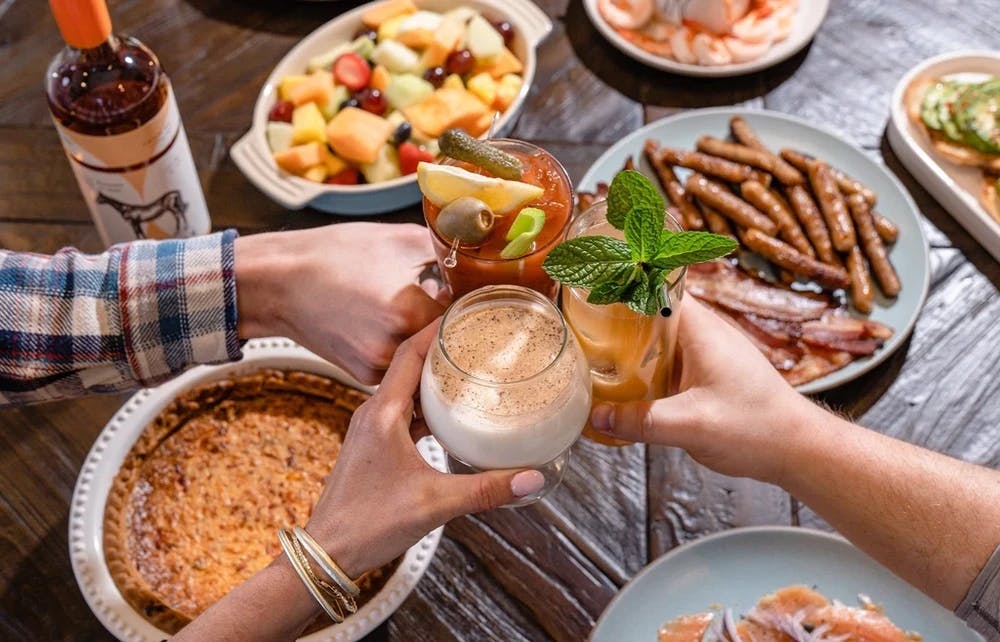 A table filled with various food and drinks, with three hands above it clinking their glasses in a toast.