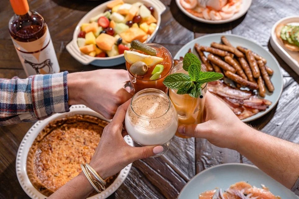 A table filled with various food and drinks, with three hands above it clinking their glasses in a toast.