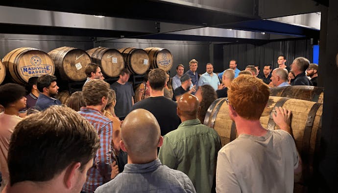 A large group of tourists gathered in a barrel storage area, attentively listening to a guide.