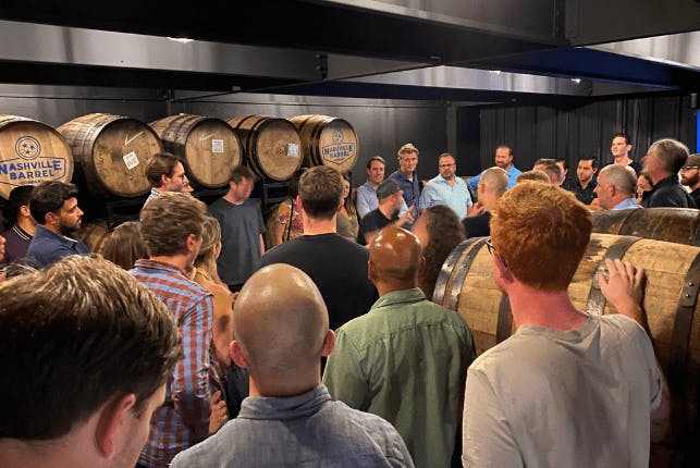 A large group of tourists gathered in a barrel storage area, attentively listening to a guide.