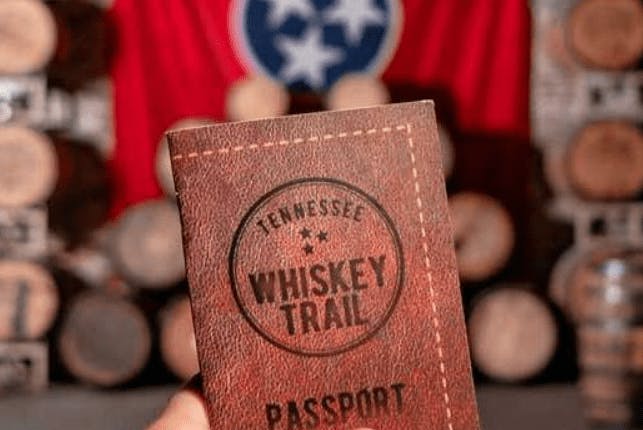 A Tennessee Whiskey Trail Passport held in focus, with a blurred background of stacked wooden whiskey barrels.