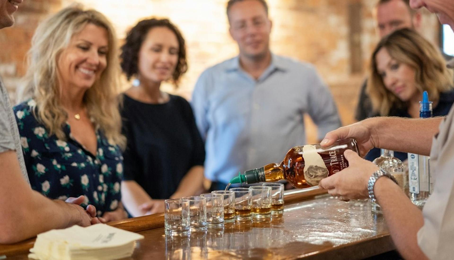 A group of on a distillery tour gathered around someone pouring Buffalo Trace into shot glasses.