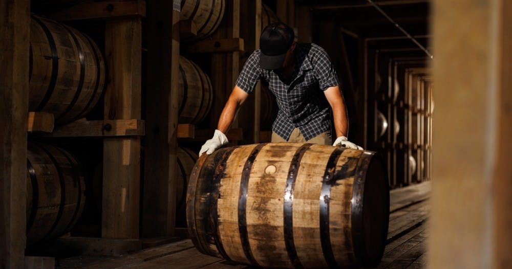 A man rolling a wooden bourbon barrel through a dark rackhouse.