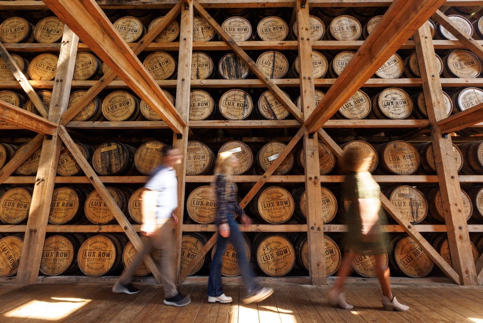 A wall stacked with wooden bourbon barrels, with three people walking in the foreground.
