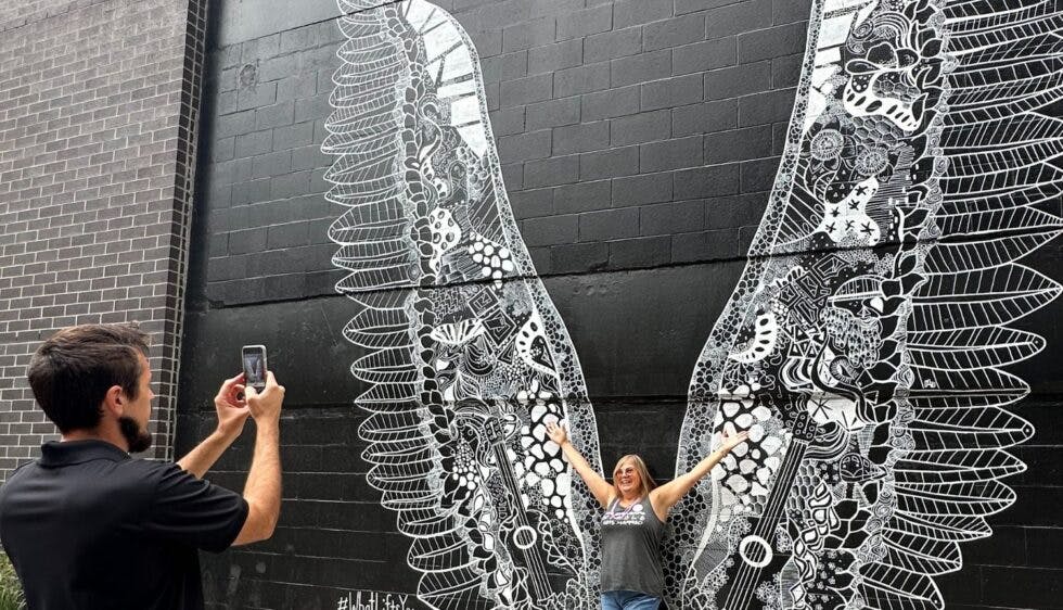 A man taking a photo of a woman posing in front of the "What Lifts You" mural in Nashville, which features giant black and white angel wings by artist Kelsey Montague, located in The Gulch neighborhood.