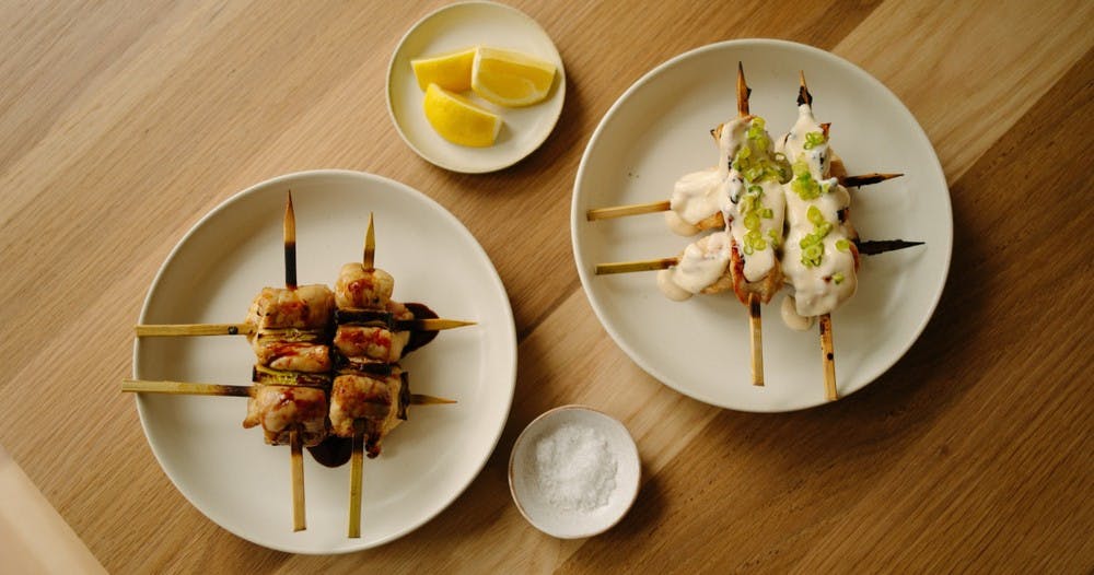 Two plates of grilled chicken skewers, accompanied by small side dishes of salt and lemon slices.