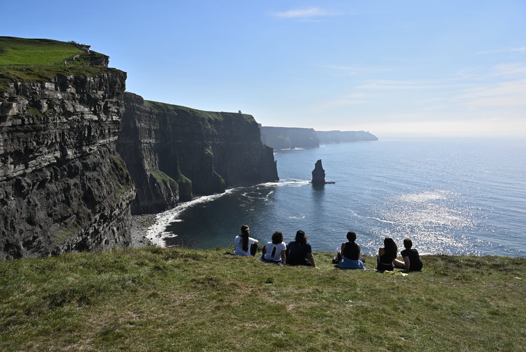 A group of tourists sitting on the grass at the Cliffs of Moher, gazing out at the scenic coastal view.