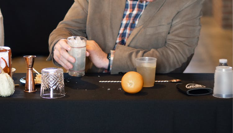 A man seated at a table, holding a glass with ingredients and tools around him, suggesting a cocktail mixing in progress.