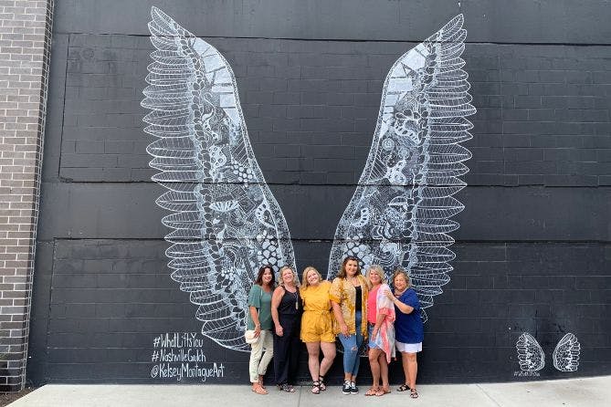 A group of women posing in front of the "What Lifts You" mural in Nashville, which features giant black and white angel wings painted by artist Kelsey Montague.