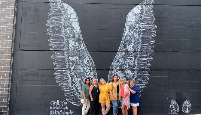 A group of women posing in front of the "What Lifts You" mural in Nashville, which features giant black and white angel wings painted by artist Kelsey Montague.
