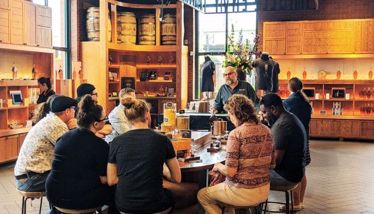 A group of people seated around a table at a distillery, participating in a bourbon tasting.