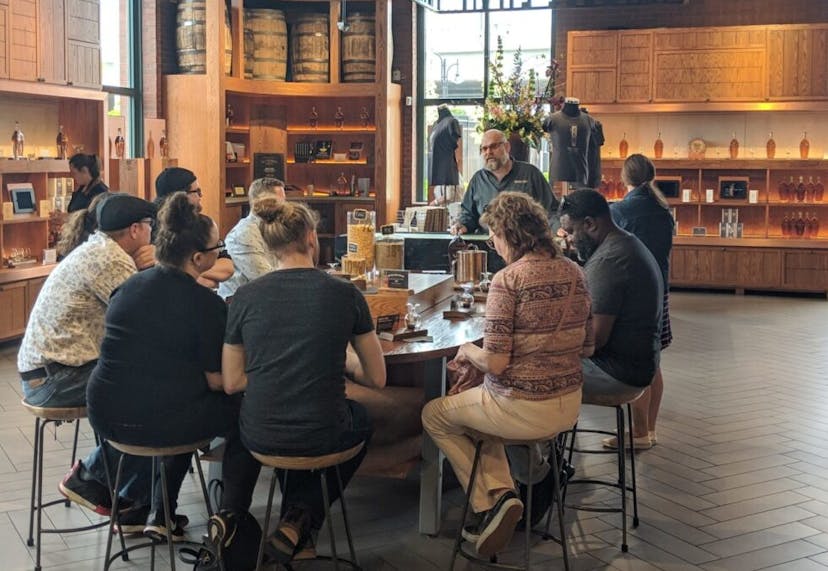 A group of people seated around an oval table at a distillery, attentively listening to a guide during a bourbon tour and tasting experience.