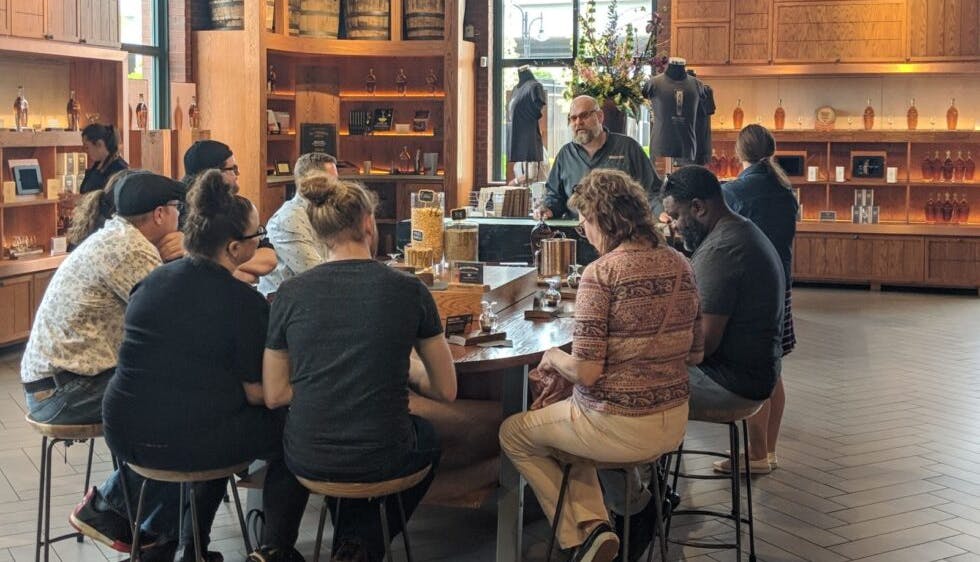 A group of people seated around an oval table at a distillery, attentively listening to a guide during a bourbon tour and tasting experience.