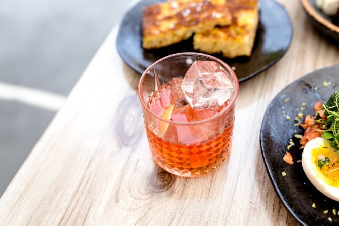 A glass of cocktail in focus, with a plate of brunch food partially visible near the edge of the frame.