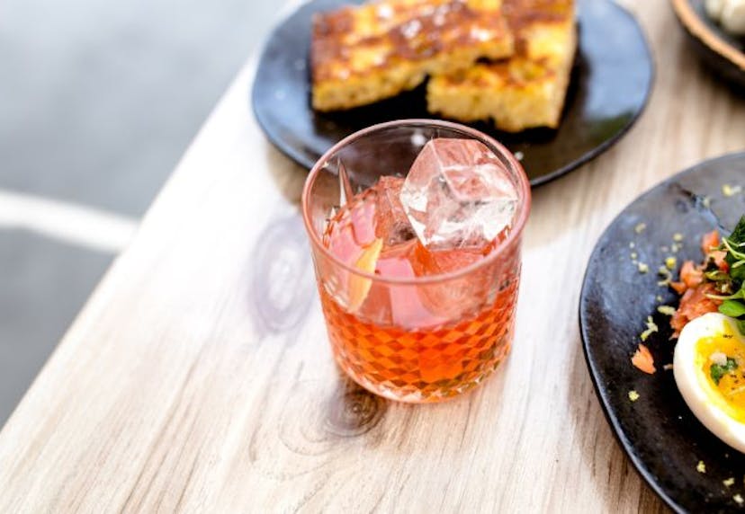 A glass of cocktail in focus, with a plate of brunch food partially visible near the edge of the frame.