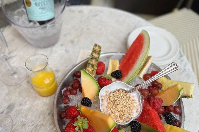 A bowl of fresh fruit with a few drinks visible in the background, suggesting a light and refreshing brunch or snack setting.