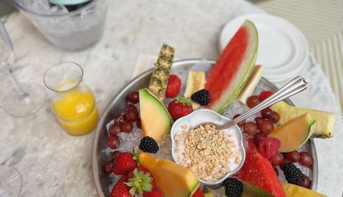 A bowl of fresh fruit with a few drinks visible in the background, suggesting a light and refreshing brunch or snack setting.