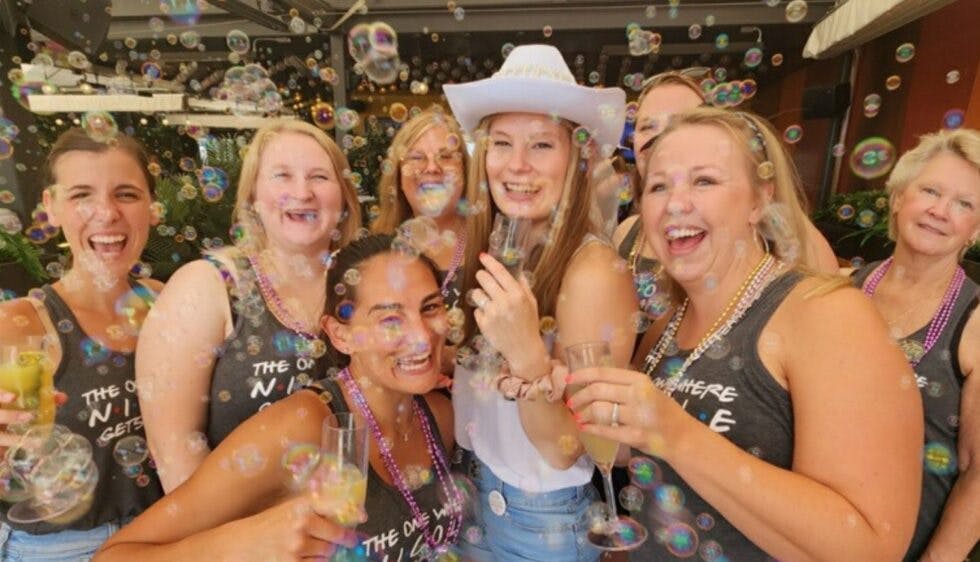 A group of women at a bachelorette party, smiling and posing for the camera while holding drinks, celebrating joyfully.