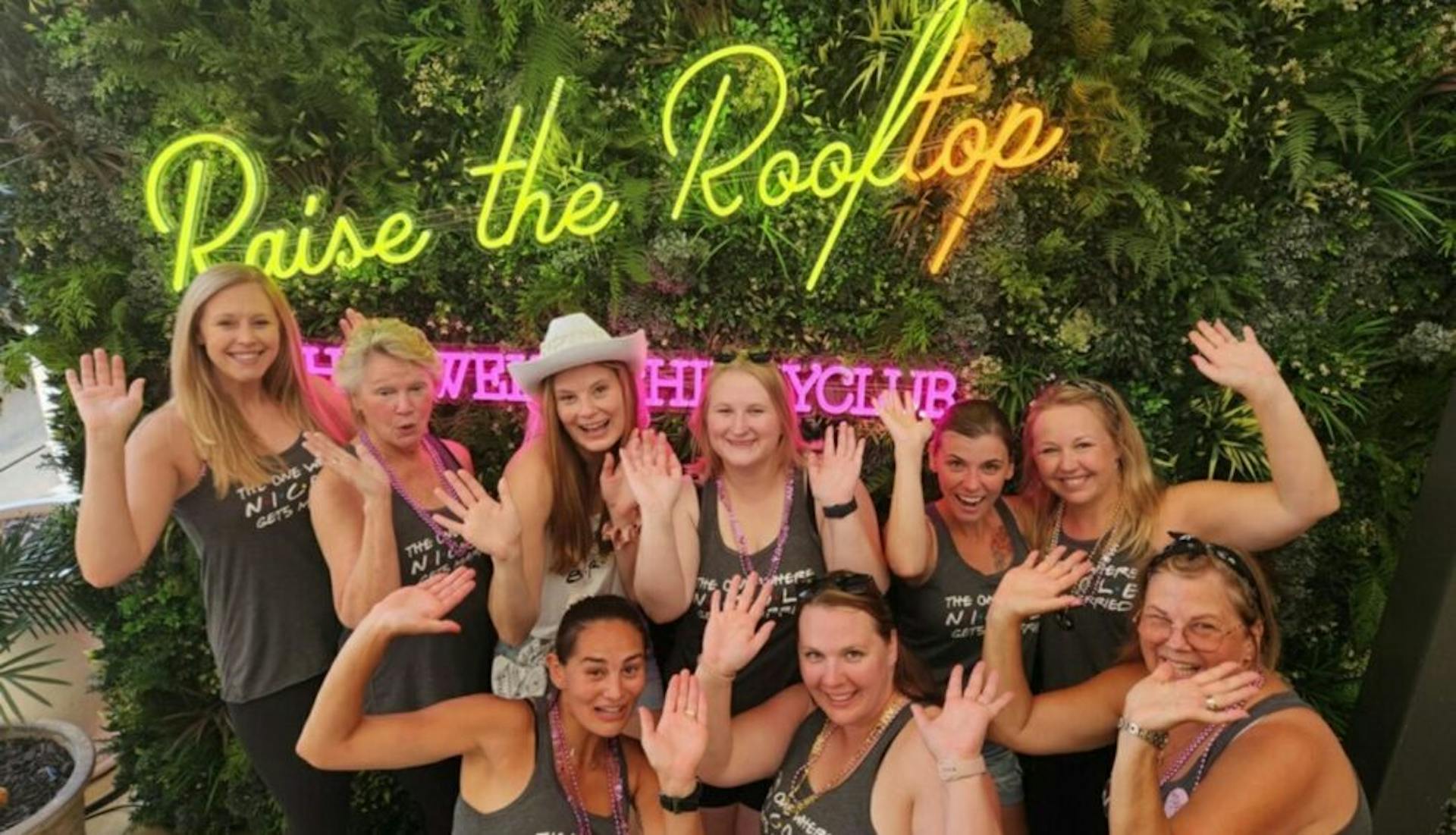 A group of women at a bachelorette party posing in front of the 'Raise the Rooftop' neon lights at The Twelve Thirty Club.