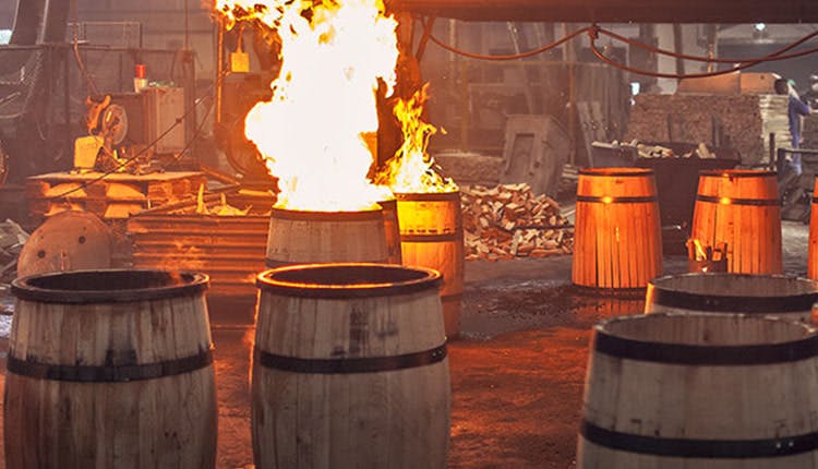 Several wooden barrels in a distillery, with some actively being charred by open flames during the barrel-making process.