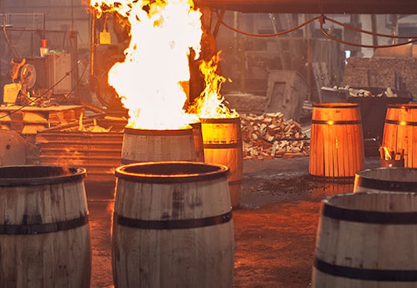 Several wooden barrels in a distillery, with some actively being charred by open flames during the barrel-making process.