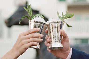 Two hands holding Mint Julep cocktails in silver cups, clinking together.