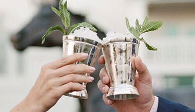 Two hands holding Mint Julep cocktails in silver cups, clinking together