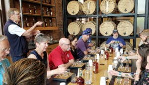 Tourists seated at a table with bourbon samples in front of them, listening as a tour guide speaks.