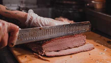 A close-up of hands slicing smoked BBQ meat on a wooden cutting board.