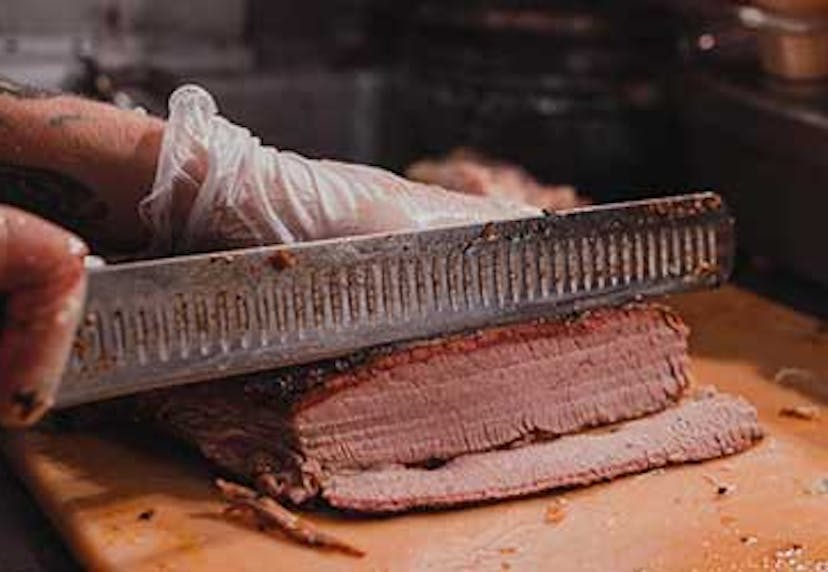 A close-up of hands slicing smoked BBQ meat on a wooden cutting board.