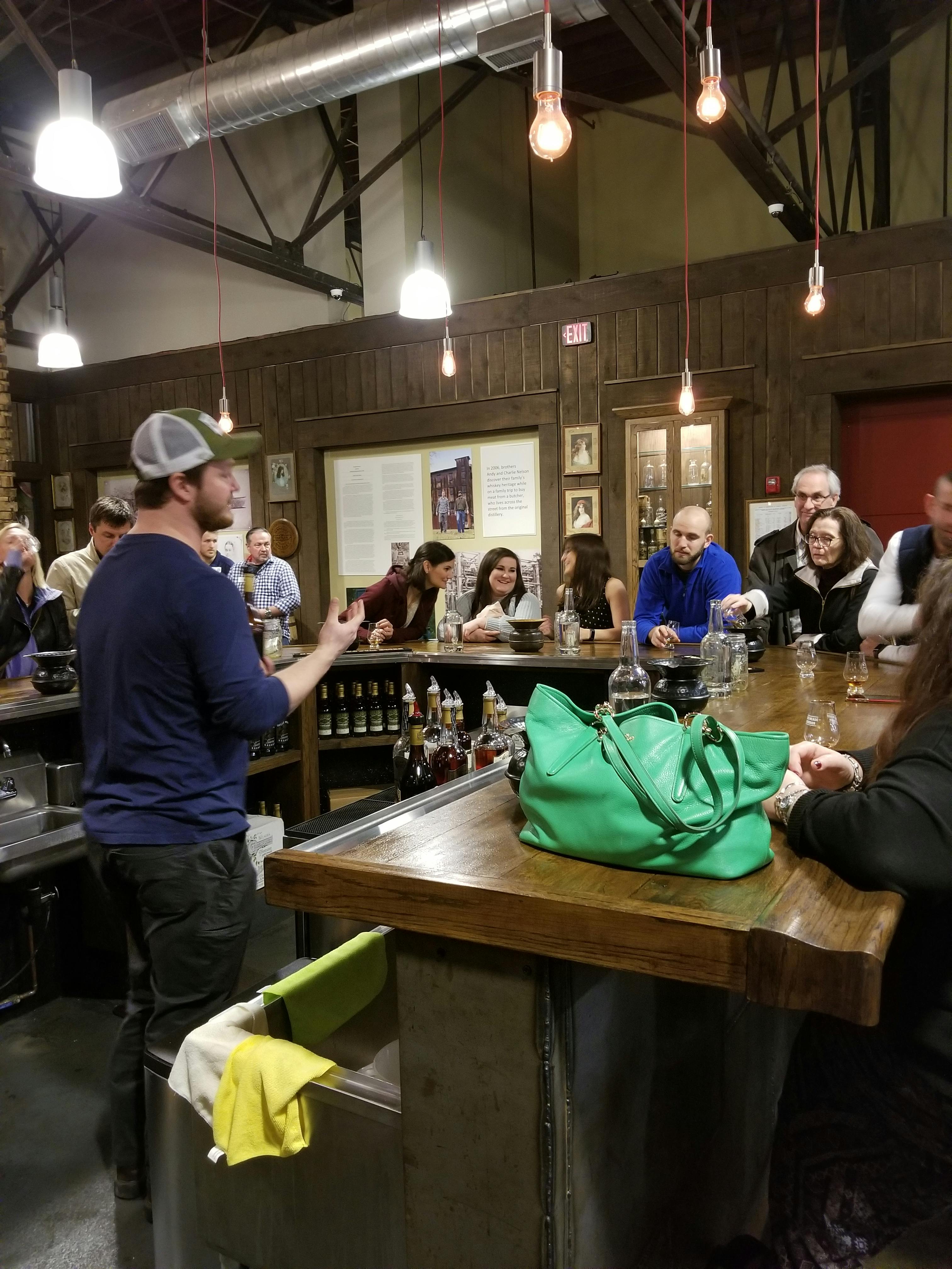 A whiskey tour at a rustic bar featuring a group of people listening to a guide behind the counter.A whiskey tasting event at a rustic bar featuring a group of people listening to a guide behind the counter.