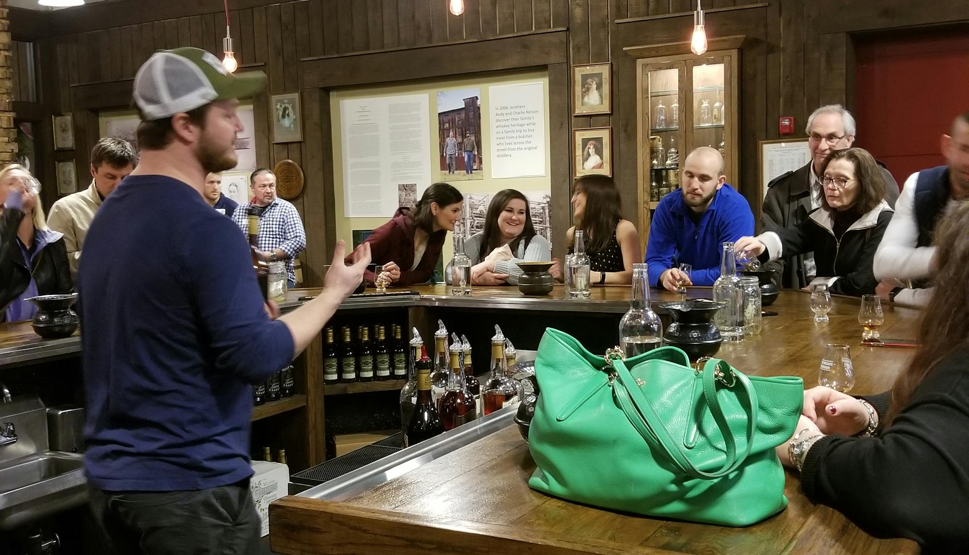 A whiskey tour at a rustic bar featuring a group of people listening to a guide behind the counter.A whiskey tasting event at a rustic bar featuring a group of people listening to a guide behind the counter.