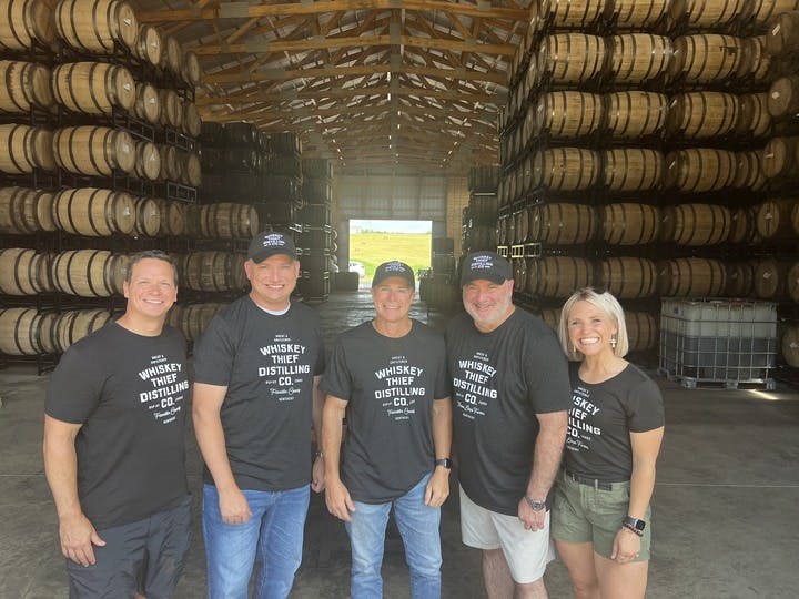 A group of people wearing Whiskey Thief Distilling Co. apparel posing for a photo inside a bourbon barrel warehouse.