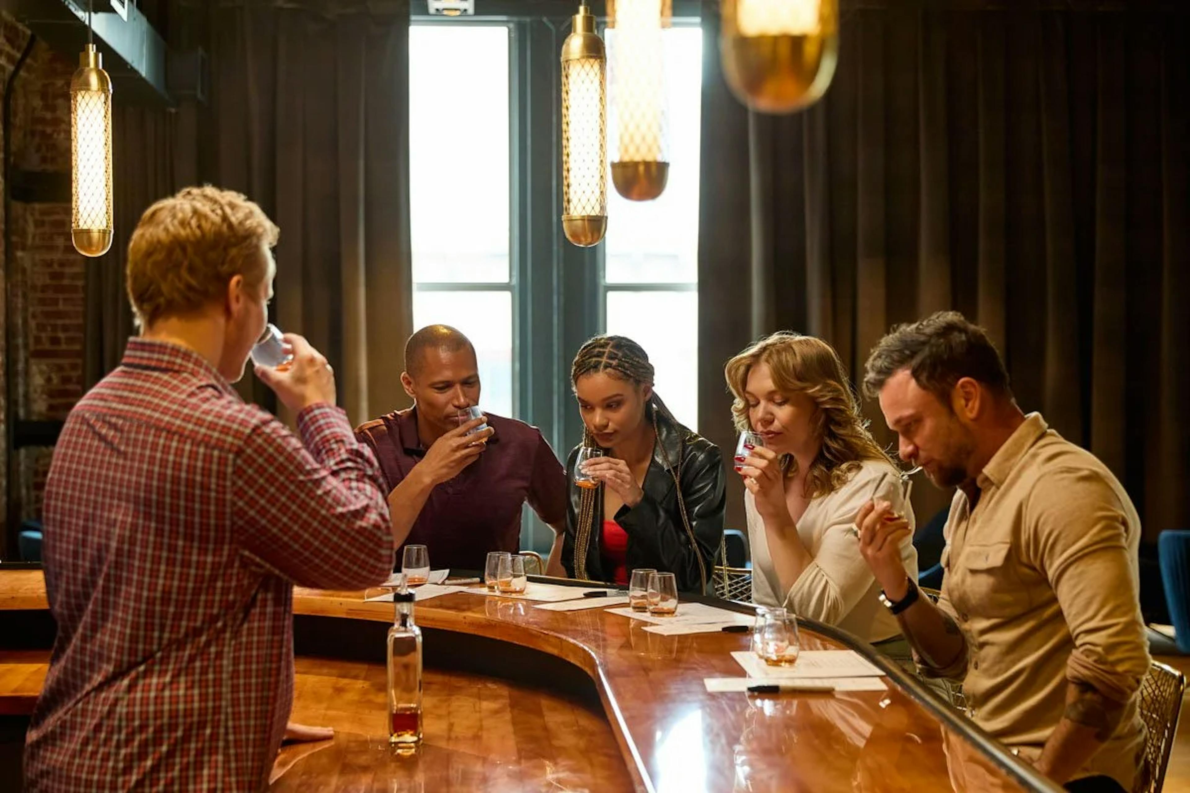 A group of men and women sit along a tasting bar and sample pappy van winkle, buffalo trace antique, king of kentucky, and william larue weller