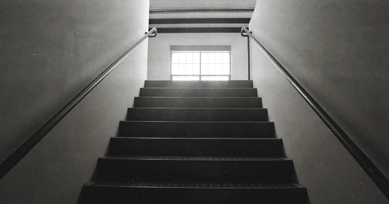 Ascending industrial staircase rising toward light, SME management consulting, the next level, contemplative black and white