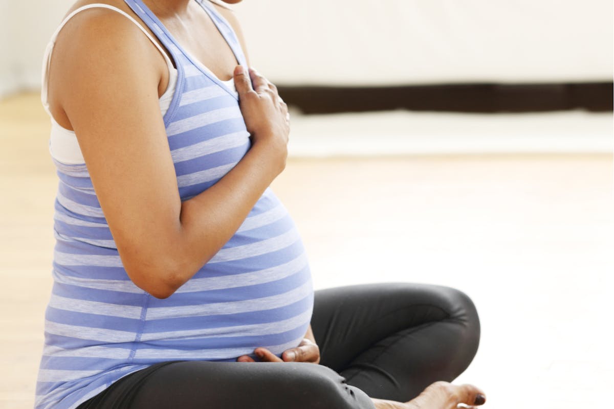 Körper einer schwangeren Frau in Yogi Sitz, jeweils eine Hand auf Herz und Bauch