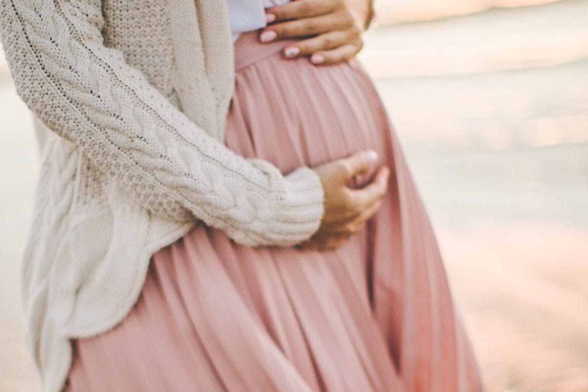 Frau hält schwangeren Bauch in ihren Händen, rosa Faltenrock, beige Strickjacke.