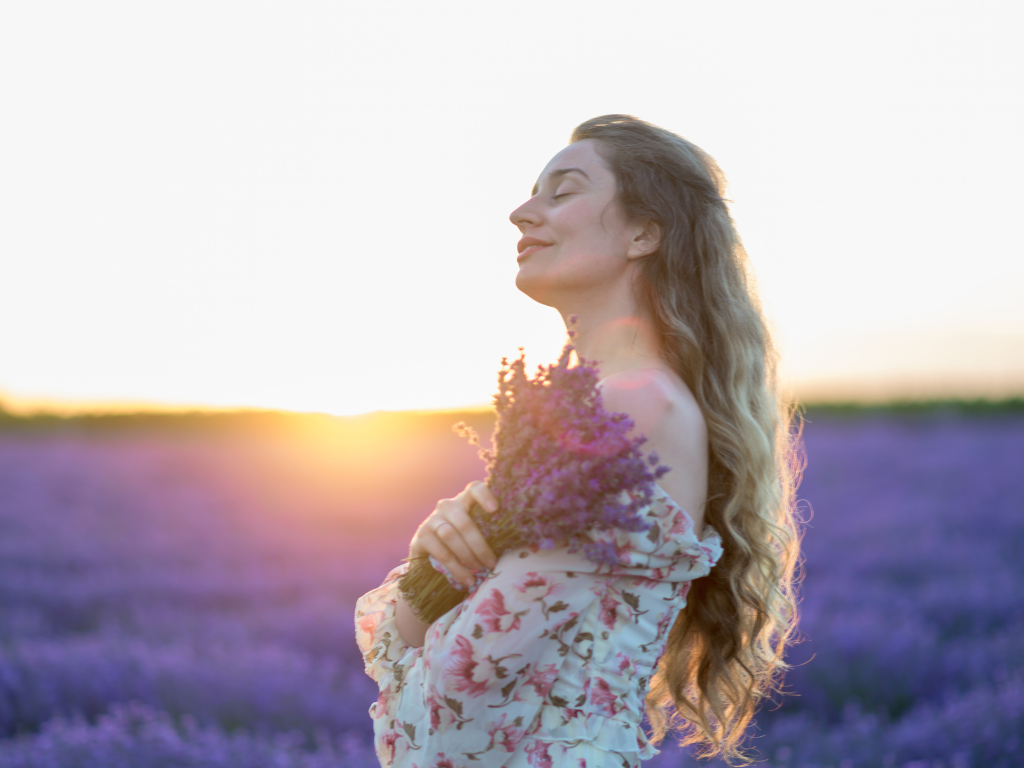 Frau mit geschlossenen Augen auf Lavendelfeld bei Sonnenuntergang mit Lavendel an ihr Herz haltend