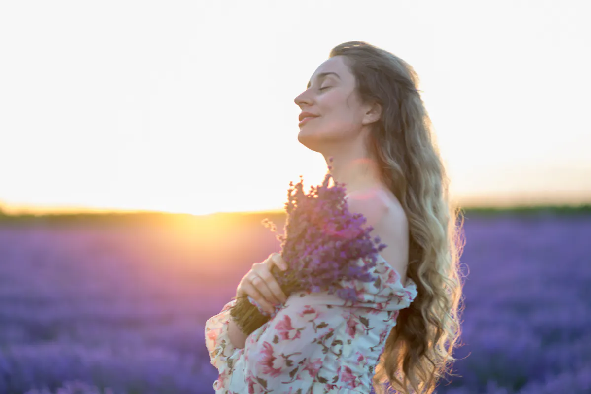 Frau mit geschlossenen Augen auf Lavendelfeld bei Sonnenuntergang mit Lavendel an ihr Herz haltend