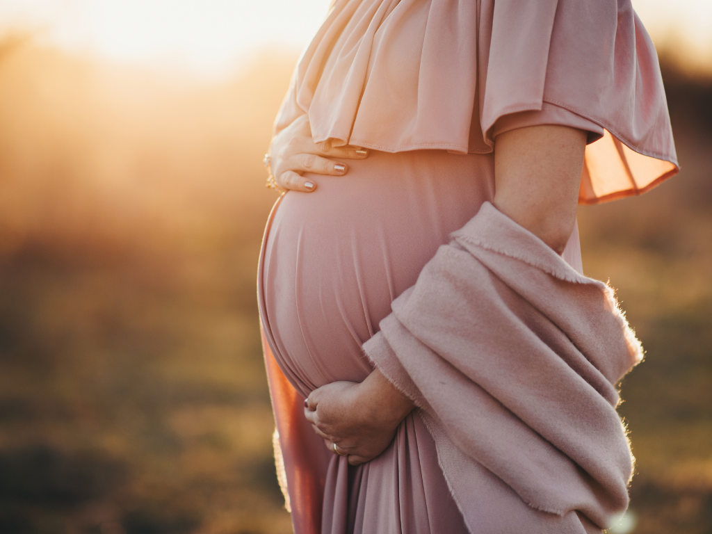 Babybauch im goldenen Sonnenlicht am Abend, Mutter trägt rosa Kleid mit Schal.