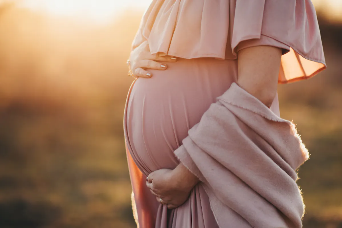 Babybauch im goldenen Sonnenlicht am Abend, Mutter trägt rosa Kleid mit Schal.