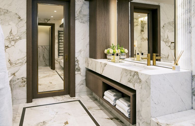 Bright marble bathroom with a neat wooden door frame, wooden storage shelves and a golden water tap in sink. 