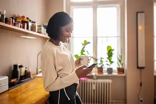 Woman in her kitchen drinking coffee while playing on her phone