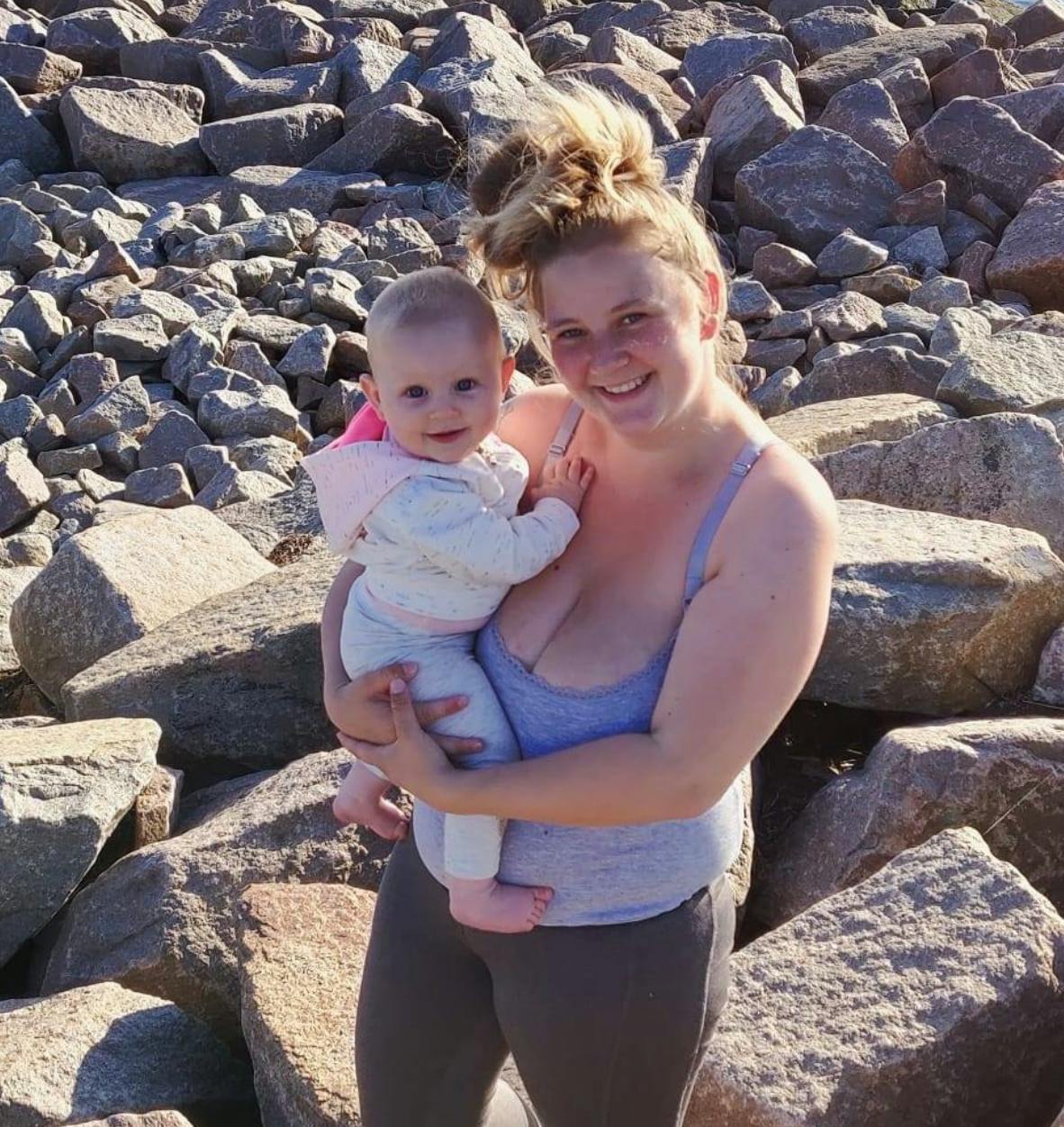 mor står med baby i armene en sommerdag med sten i baggrunden