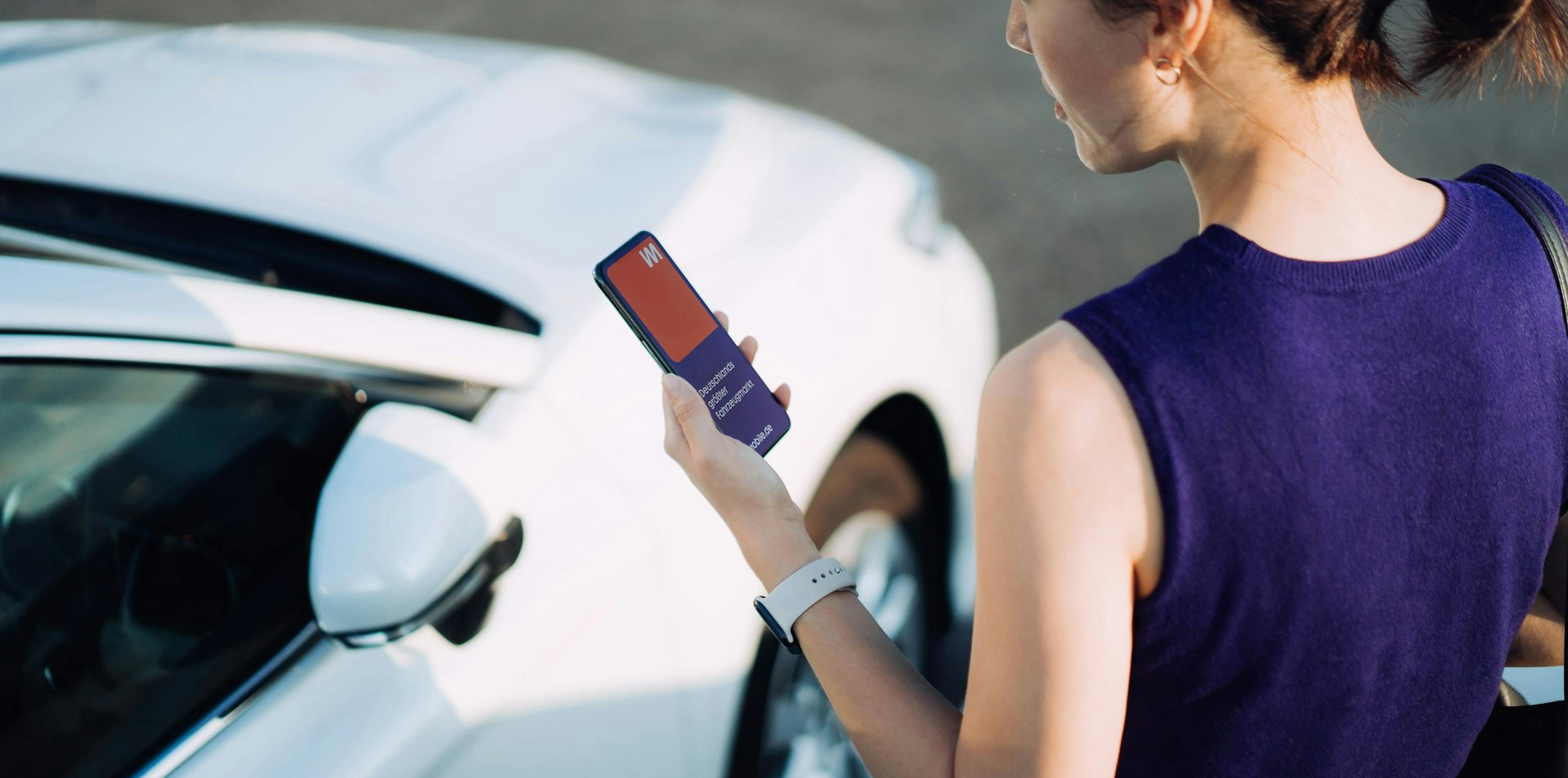 Frau schaut auf ihr Handy mit der mobile.de App, vor einem weißen Auto