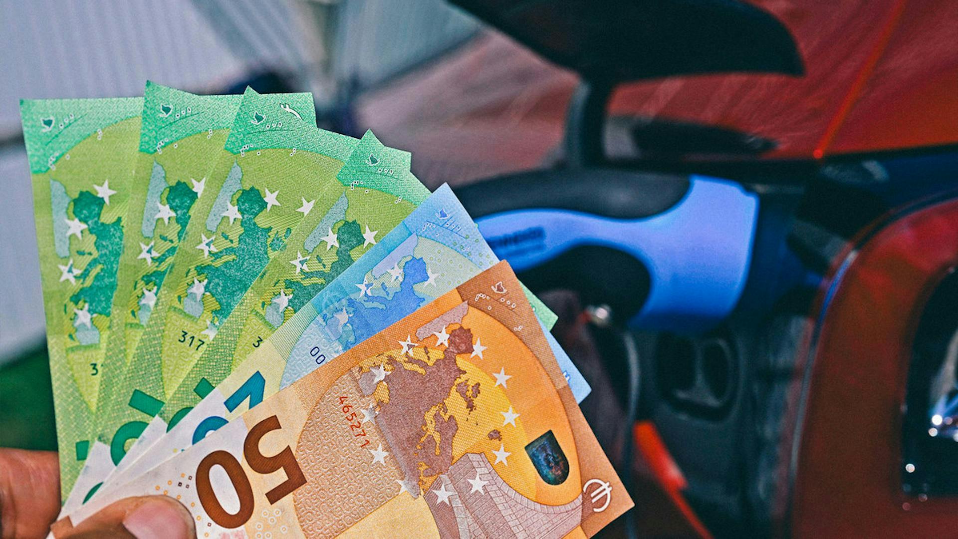 Eine Hand hält Geldscheine hoch, im Hintergrund steht ein E-Auto