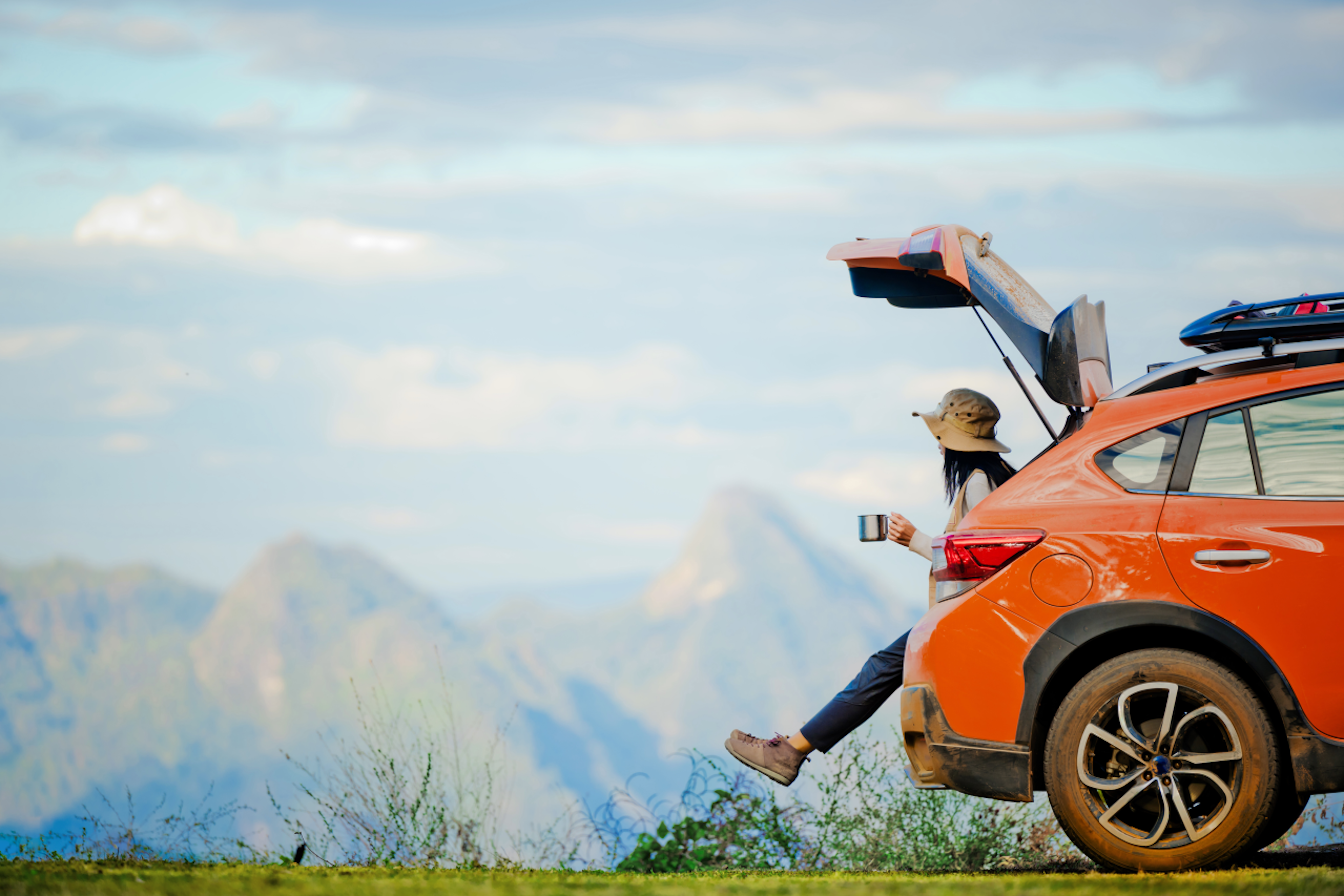Frau sitz in der geöffneten Motorhaube ihres Wagens und schaut mit einer Kaffeetasse in der Hand auf die Berge.