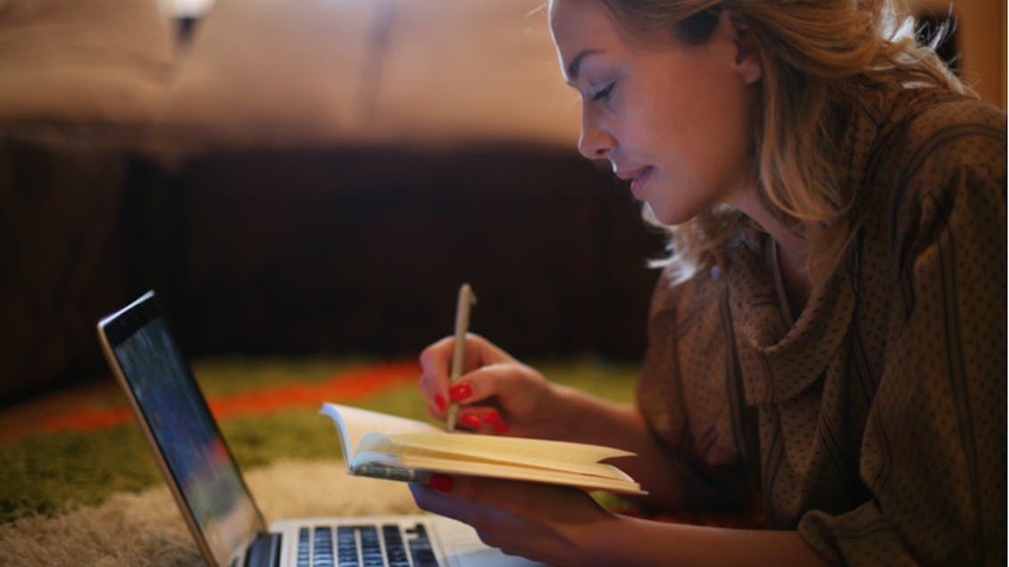 A woman looking at her computer and writing something on a book 