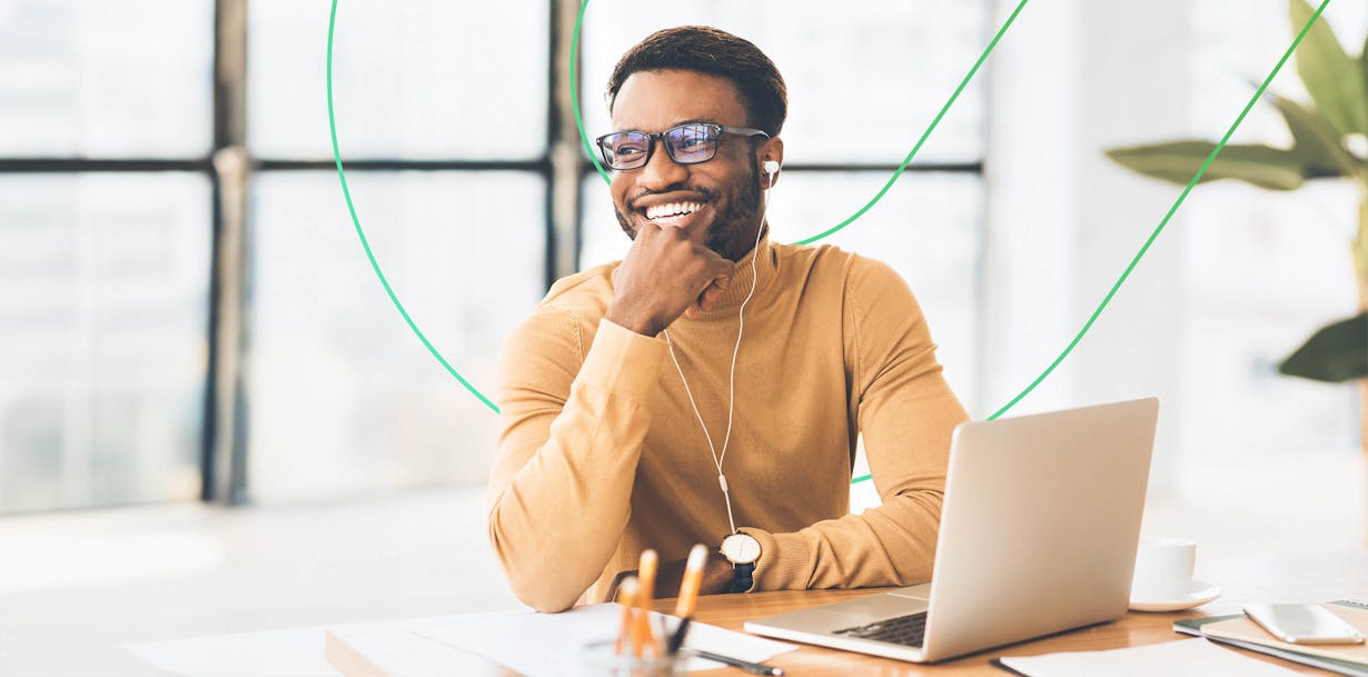 Black man smiling with laptop at a desk with a window behind him
