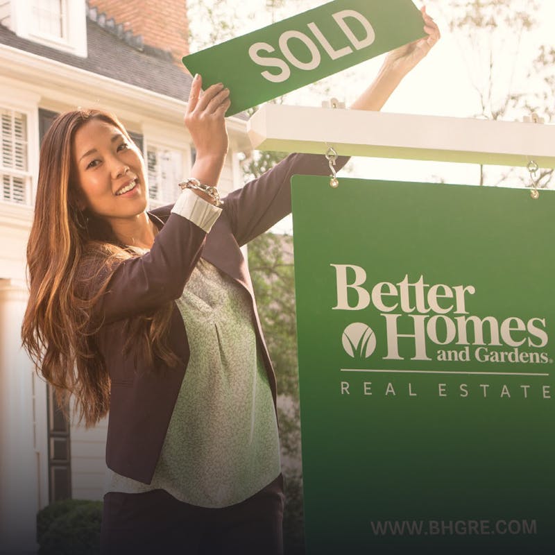 A saleswoman holding a sold sign over a Better Homes and Gardens banner